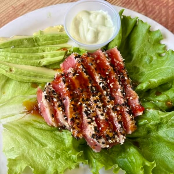 Seared tuna with sesame seeds, avocado slices, and a side of creamy sauce on lettuce.
