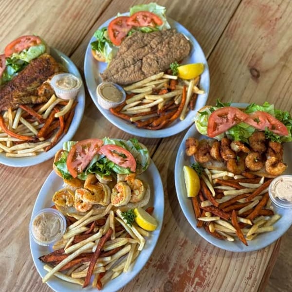 Four plates of seafood po'boys with fries, sweet potato fries, and dipping sauce on a wooden table.