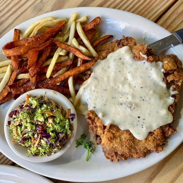 Fried chicken steak with gravy, fries, sweet potato fries, and broccoli slaw on a white plate.