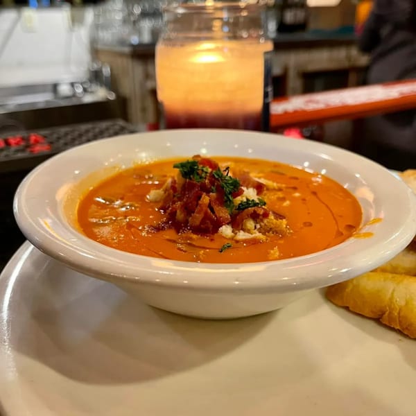 A bowl of orange seafood bisque, topped with bacon bits and herbs, next to bread.