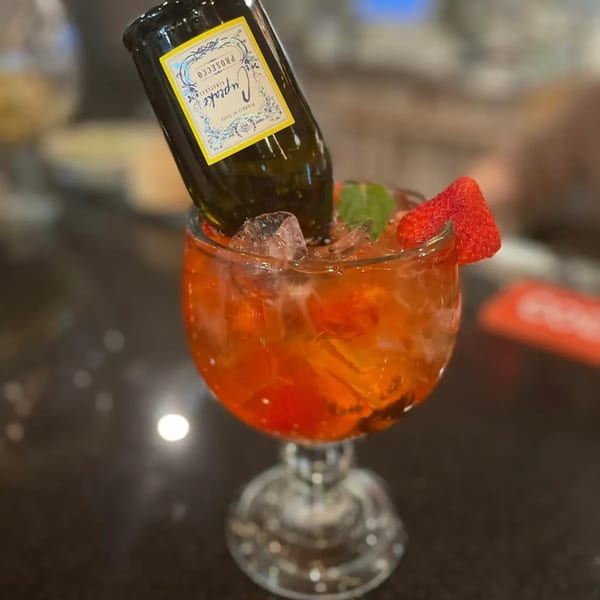 A vibrant orange cocktail with ice, garnished with a strawberry and mint, featuring an inverted mini Prosecco bottle.