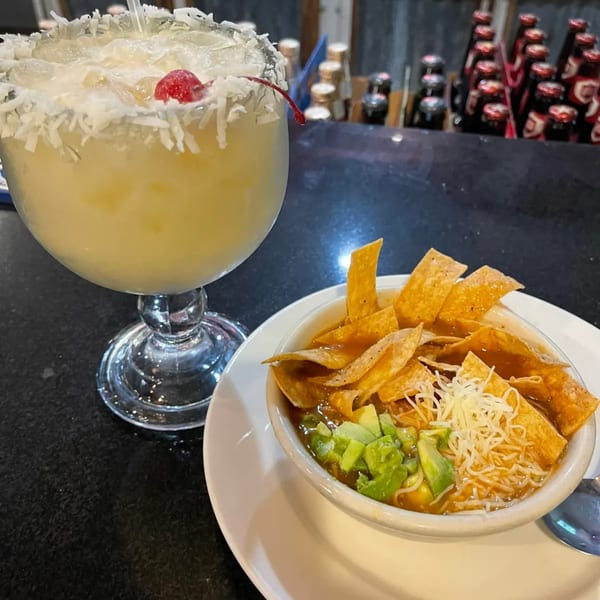 A coconut margarita with a cherry and a bowl of tortilla soup with avocado and cheese.