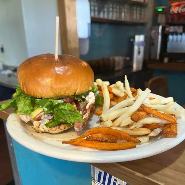 A grilled chicken sandwich with lettuce, avocado, and bacon, served with fries and sweet potato fries.